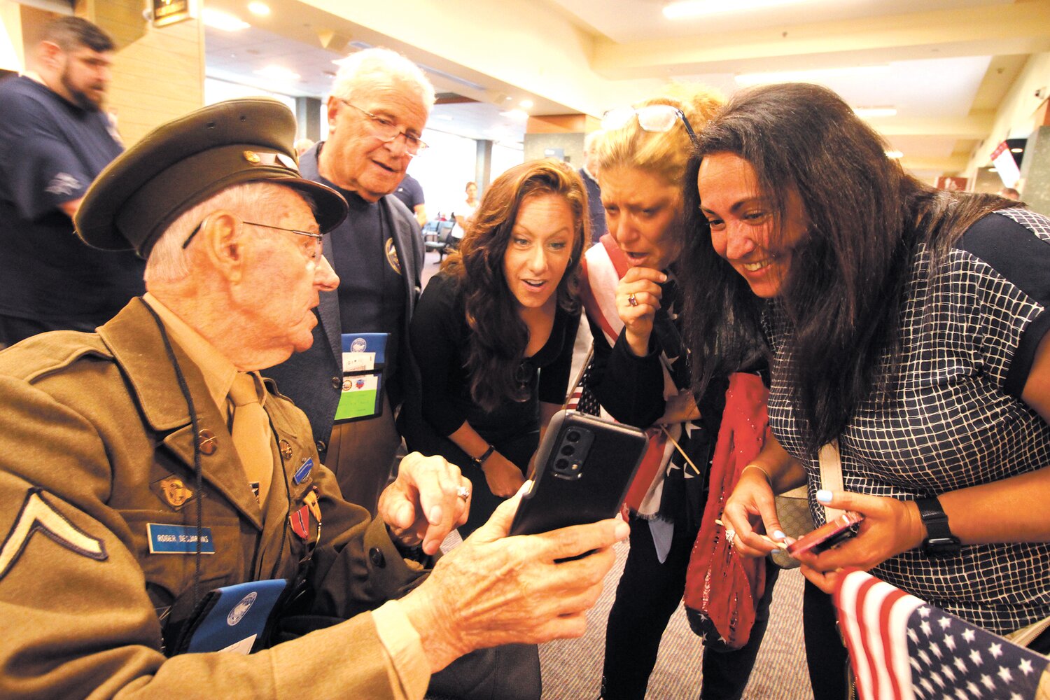 98 AND HIGH TECH: WWII Army veteran Roger Desjardins of North Providence, the oldest member of Honor Flight Freedom that left from Green Airport Monday, pulled out his cell phone to accompany stories he was recounting. Behind him is North Providence Mayor Charles Lombardi who was Desjardins’ guardian for the flight. (Cranston Herald photos by John Howell)